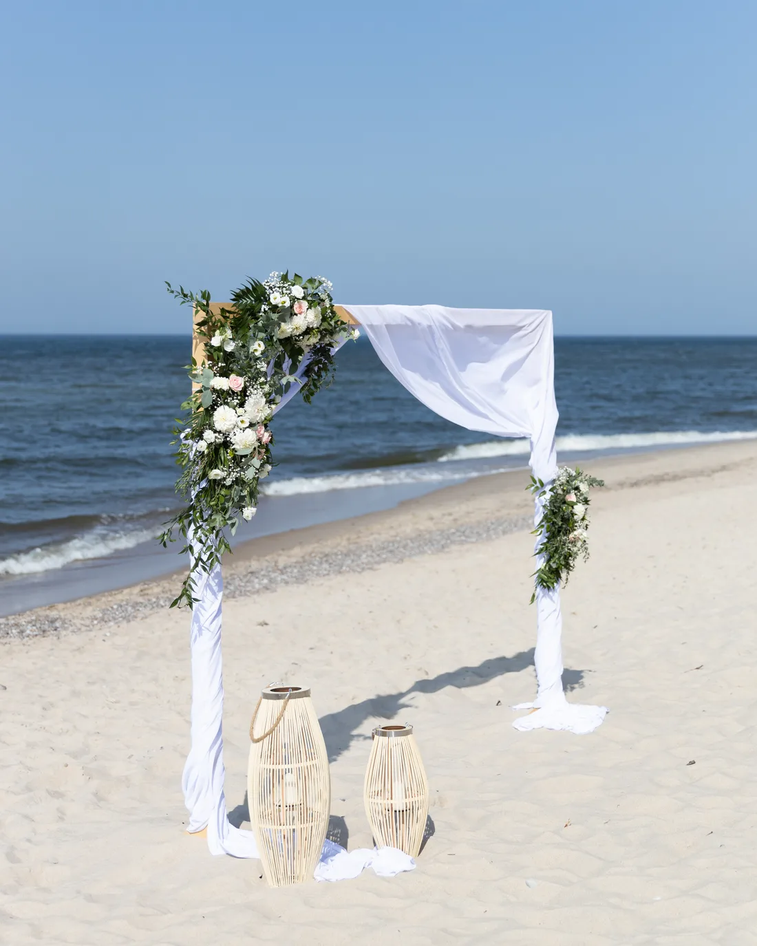 Łuk kwiatowy z białą draperią i lampionami na plaży nad morzem